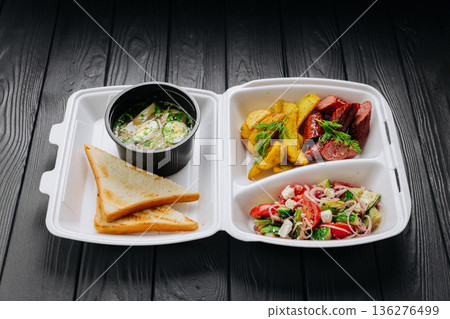 Takeout box with chicken broth soup, toast, fresh salad, potato wedges and sausage 136276499