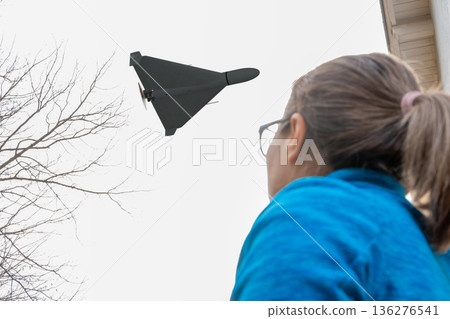 A woman outdoors observes a flying military drone above houses and trees. 136276541