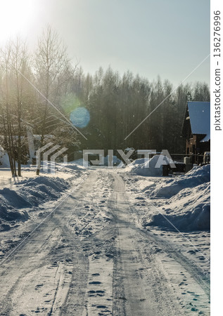 Sunny winter day on a snowy country road with tire tracks and lens flare, leading towards a forest and rural homes. 136276996