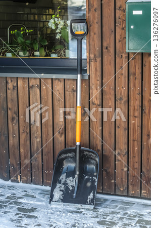 Snow shovel leaning against a wooden house wall on a snowy winter day. Winter chore essential. 136276997