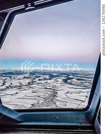 Pilots view from an airplane cockpit of a snowy winter landscape with a village and patchwork fields below. 136276998
