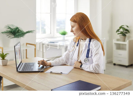 Female doctor sitting in front of laptop, writing report or prescription after patient examination. Female doctor sitting in front of laptop, writing report or prescription after patient examination. 136277349