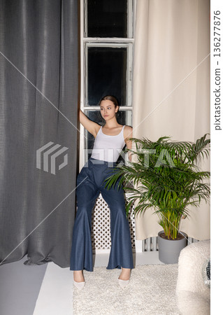 Young woman wearing bell-bottom jeans standing by window 136277876
