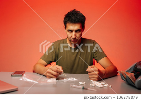 Serious drug dealer male preparing to snort cocaine with rolled banknote sitting at table with pills and syringe on red background, looking at camera, representing drug addiction and substance abuse. 136278879