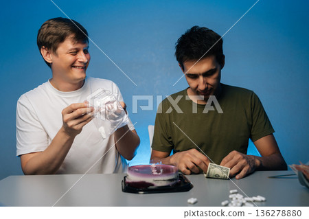 Cheerful male friends sitting at table, one snorting white powder from plastic bag while other arranging rolled dollar bills during illicit substance interaction. Concept of party with substance abuse 136278880
