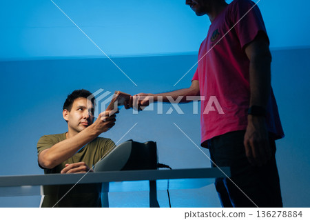 Police officer receiving bribe money from businessman in dark room, using counterfeit money detector to check banknotes authenticity, under blue light, low-angle view, close-up, studio shot. Police officer receiving bribe money from businessman in dark room, using counterfeit money detector to check banknotes authenticity, under blue light, low-angle view, close-up, studio shot. 136278884