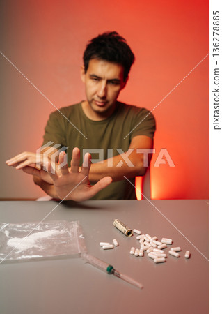 Vertical portrait of depressed man showing stop sign with hand while refusing drugs, cocaine, pills rolled banknote, sitting at table with syringe on red background. Concept of substance abuse. 136278885