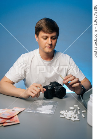 Vertical portrait of photographer preparing drugs with rolled banknote sitting at desk with camera, struggling with addiction, studio shot portrait on isolated background. Concept of substance abuse. Vertical portrait of photographer preparing drugs with rolled banknote sitting at desk with camera, struggling with addiction, studio shot portrait on isolated background. Concept of substance abuse. 136278886