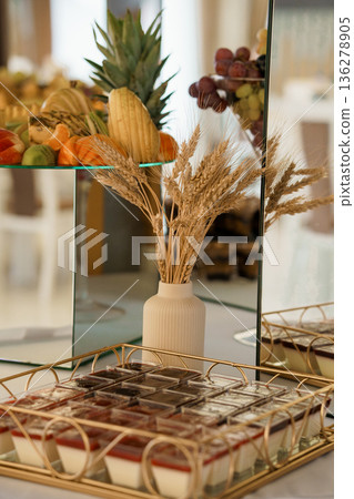 Elegant dessert buffet display with golden wheat arrangement, pineapple, grapes, and colorful fruit platter on mirrored tiered stand Elegant dessert buffet display with golden wheat arrangement, pineapple, grapes, and colorful fruit platter on mirrored tiered stand 136278905