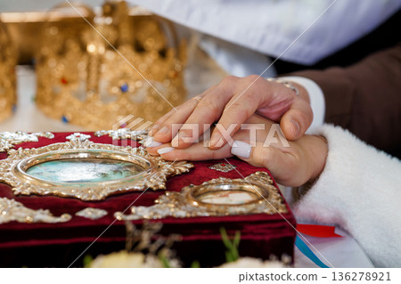 Elegant Wedding Ceremony Hands Holding Over Ornate Religious Icon with Gold Crown and Red Velvet Background 136278921