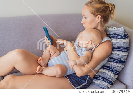 Baby sitting on mother lap while she looks at her smartphone, emotionally disconnected. Symbolic scene showing impact of screen distraction on infants and unmet emotional needs. Digital parenting and Baby sitting on mother lap while she looks at her smartphone, emotionally disconnected. Symbolic scene showing impact of screen distraction on infants and unmet emotional needs. Digital parenting and 136279249