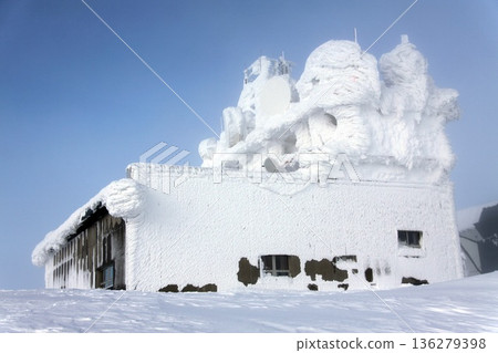 The image depicts a remote, utility building with telecommunications antennas and microwave dishes completely buried in snow and ice. 136279398