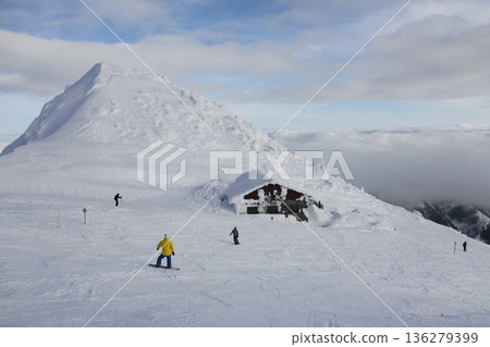 a snow-buried alpine mountain hut on an open ski slope, with snowboarders and skiers passing by 136279399