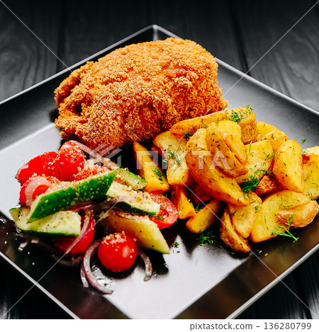 Breaded chicken cutlet with roasted potatoes and fresh salad on black plate, dark table Breaded chicken cutlet with roasted potatoes and fresh salad on black plate, dark table 136280799