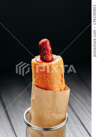 Creative hot dog in baguette with ketchup in paper bag on dark wooden background 136280801