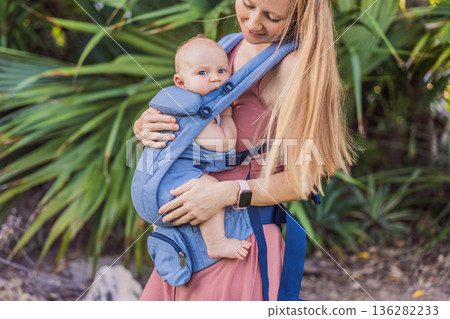 Mother using ergonomic baby carrier during a walk with her infant outdoors. Comfortable babywearing, active motherhood lifestyle, bonding, modern parenting, hands free care and everyday family routine Mother using ergonomic baby carrier during a walk with her infant outdoors. Comfortable babywearing, active motherhood lifestyle, bonding, modern parenting, hands free care and everyday family routine 136282233
