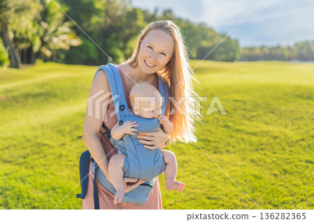 Mother using ergonomic baby carrier during a walk with her infant outdoors. Comfortable babywearing, active motherhood lifestyle, bonding, modern parenting, hands free care and everyday family routine Mother using ergonomic baby carrier during a walk with her infant outdoors. Comfortable babywearing, active motherhood lifestyle, bonding, modern parenting, hands free care and everyday family routine 136282365