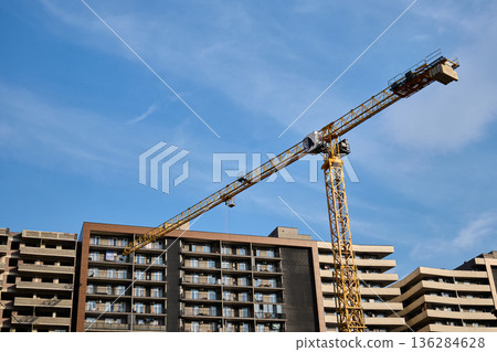 A large yellow tower crane stands over a multi-story residential building under a clear blue sky A large yellow tower crane stands over a multi-story residential building under a clear blue sky 136284628