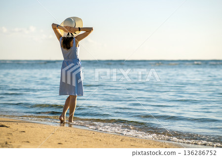 Young woman in straw hat and a dress walking alone on empty sand beach at sea shore. Lonely tourist girl looking at horizon over calm ocean surface on vacation trip. 136286752