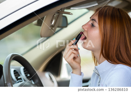 Closeup of a young redhead woman driver correcting her makeup with dark red lipstick looking in car rearview mirror behind steering wheel of a vehicle. 136286810