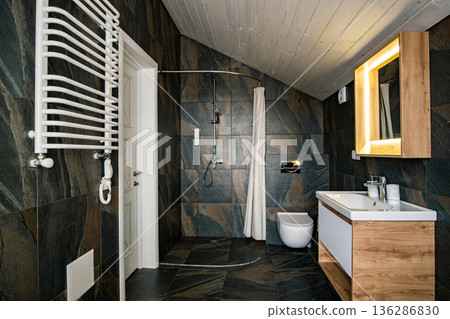 Interior of modern stylish bathroom with black tiled walls, curtain shower place and wooden furniture with wash basin and big illuminated mirror. 136286830