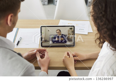 Company employees having online video call with colleague using tablet sitting in office. Company employees having online video call with colleague using tablet sitting in office. 136286997