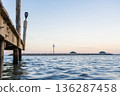 Wooden Pier Over Calm Sea with Distant Lighthouse at Sunset 136287458