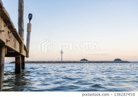 Wooden Pier Over Calm Sea with Distant Lighthouse at Sunset Wooden Pier Over Calm Sea with Distant Lighthouse at Sunset 136287458