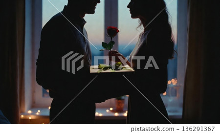 Silhouette couple exchanging rose and gift by window at dusk Silhouette couple exchanging rose and gift by window at dusk 136291367