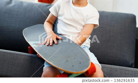 Child holds skateboard while sitting on sofa in a bright indoor space during the day Child holds skateboard while sitting on sofa in a bright indoor space during the day 136291635