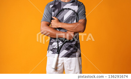Tennis player stands with racket and smiles in studio against orange background 136291674