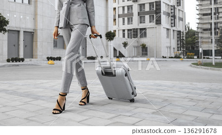 Woman walks in urban area with suitcase during daylight near modern buildings 136291678