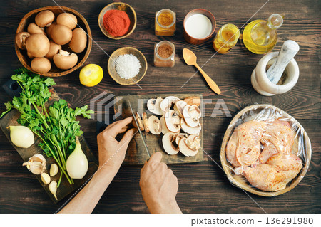 Top view of chef hands slicing fresh mushroom on wooden board with marinated chicken nearby 136291980