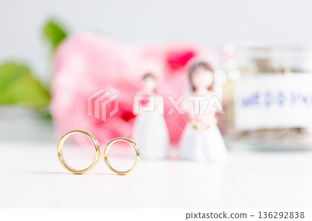 Inclusive wedding commitment concept celebrating equality, love and shared planning with pastel copy space; two gold rings in focus, blurred bride figurines, pink flowers and coin jar. celebrating Inclusive wedding commitment concept celebrating equality, love and shared planning with pastel copy space; two gold rings in focus, blurred bride figurines, pink flowers and coin jar. celebrating 136292838