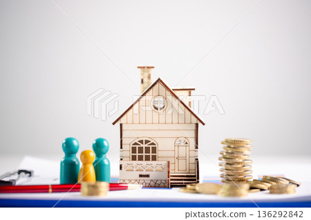 Homeownership planning and household budgeting concept for mortgage and savings, featuring miniature wooden house with coin stacks, colorful family figures and pen on white clipboard. mortgage saving 136292842