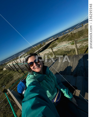 Smiling solo woman taking selfie on wooden boardwalk near sandy beach and Atlantic Ocean. Camino de Santiago, solo travel, pilgrimage, freedom Smiling solo woman taking selfie on wooden boardwalk near sandy beach and Atlantic Ocean. Camino de Santiago, solo travel, pilgrimage, freedom 136295548
