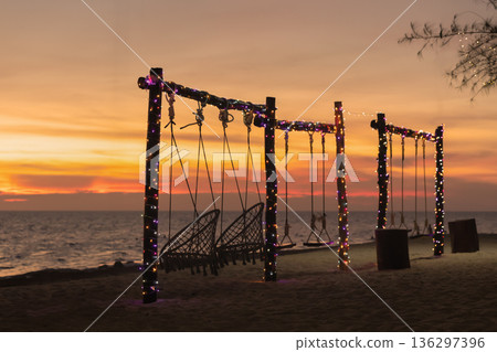 Swings are set up on the beach as the sun sets in the background. Lights hang from the swing posts, and the waves of the sea are visible nearby. High quality photo Swings are set up on the beach as the sun sets in the background. Lights hang from the swing posts, and the waves of the sea are visible nearby. High quality photo 136297396