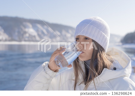 A woman holds a glass of clear water while standing outdoors in winter near a natural spring. Snow covers the ground, and sunlight brightens the scene. High quality photo A woman holds a glass of clear water while standing outdoors in winter near a natural spring. Snow covers the ground, and sunlight brightens the scene. High quality photo 136298624