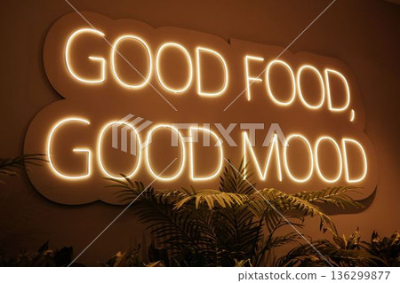 Warm neon wall sign reading Good Food Good Mood glowing in a cozy interior with plants, creating a relaxed atmosphere linked to dining, comfort, and positive lifestyle concepts 136299877