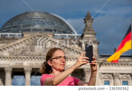 Berlin, germany, august 12, 2023. Woman tourist using a smartphone for capturing the reichstag building and fluttering german flag, experiencing travel in berlin Berlin, germany, august 12, 2023. Woman tourist using a smartphone for capturing the reichstag building and fluttering german flag, experiencing travel in berlin 136300362