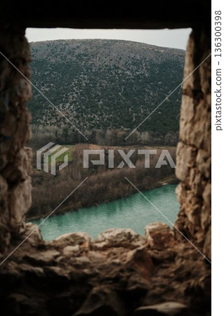 Turquoise Neretva river flowing beneath forested hills and open fields, viewed through rough stone walls of Pocitelj Fortress in a calm natural landscape Turquoise Neretva river flowing beneath forested hills and open fields, viewed through rough stone walls of Pocitelj Fortress in a calm natural landscape 136300398