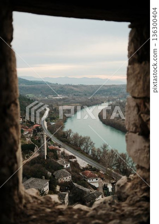 Winding Neretva river and rural road seen from a narrow stone opening inside Pocitelj Fortress, flowing through a quiet valley with villages and distant mountains Winding Neretva river and rural road seen from a narrow stone opening inside Pocitelj Fortress, flowing through a quiet valley with villages and distant mountains 136300404