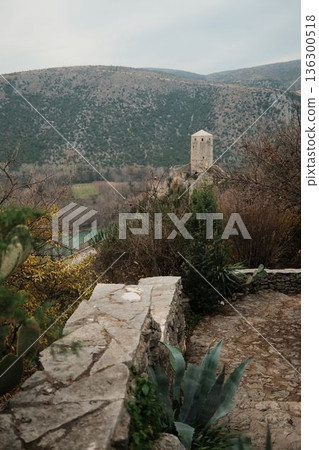 Stone path and old wall lead toward a medieval watchtower in Pocitelj Fortress surrounded by hills and river landscape. The scene reflects historic architecture blended with rugged Balkan nature 136300518