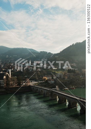 Elevated view shows the historic stone bridge crossing the Drina River with Visegrad town and hills in the background, Bosnia and Herzegovina. Light haze adds depth and atmosphere 136300522