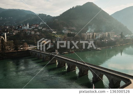Elevated view shows the historic stone bridge crossing the Drina River with Visegrad town and hills in the background, Bosnia and Herzegovina. Light haze adds depth and atmosphere Elevated view shows the historic stone bridge crossing the Drina River with Visegrad town and hills in the background, Bosnia and Herzegovina. Light haze adds depth and atmosphere 136300526