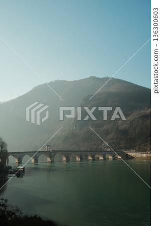 Stone bridge crosses the Drina River with forested hills rising above Visegrad, Bosnia and Herzegovina. Soft morning light creates a calm and atmospheric mountain landscape Stone bridge crosses the Drina River with forested hills rising above Visegrad, Bosnia and Herzegovina. Soft morning light creates a calm and atmospheric mountain landscape 136300603