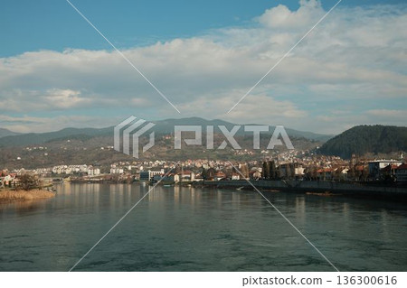Wide view of the Drina River flowing past Visegrad with hills and residential buildings along the riverbank. Calm water and open sky emphasize the natural rhythm of the town 136300616