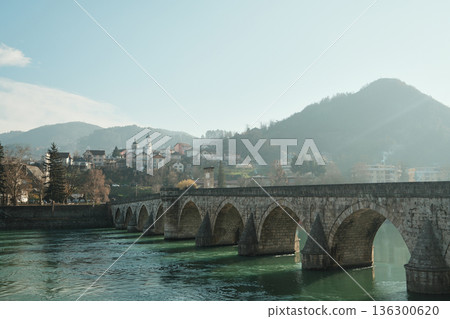 Mehmed Pasha Sokolovic Bridge stretches across the Drina River with Visegrad town and hills in the background. Soft light and mist create a peaceful historic landscape Mehmed Pasha Sokolovic Bridge stretches across the Drina River with Visegrad town and hills in the background. Soft light and mist create a peaceful historic landscape 136300620
