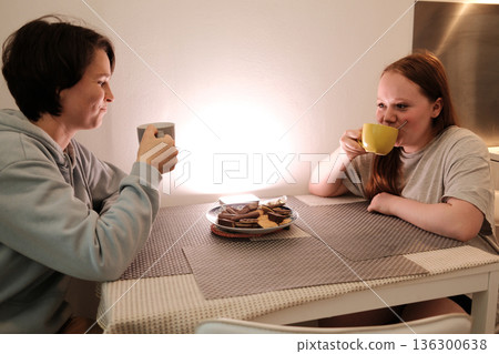 Two friends sit at a kitchen table, drinking tea and sharing cookies in a warm atmosphere. The concept emphasizes comfort, hospitality and friendly connection Two friends sit at a kitchen table, drinking tea and sharing cookies in a warm atmosphere. The concept emphasizes comfort, hospitality and friendly connection 136300638