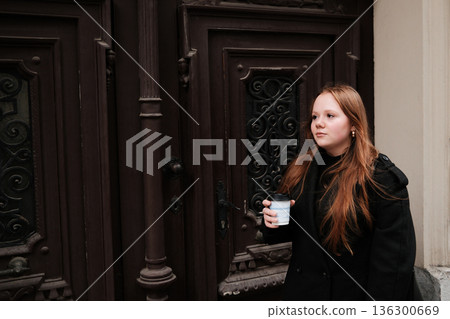 A young woman walks along the street holding a coffee cup near an ornate vintage door. The concept conveys everyday life, movement and an urban cultural atmosphere A young woman walks along the street holding a coffee cup near an ornate vintage door. The concept conveys everyday life, movement and an urban cultural atmosphere 136300669